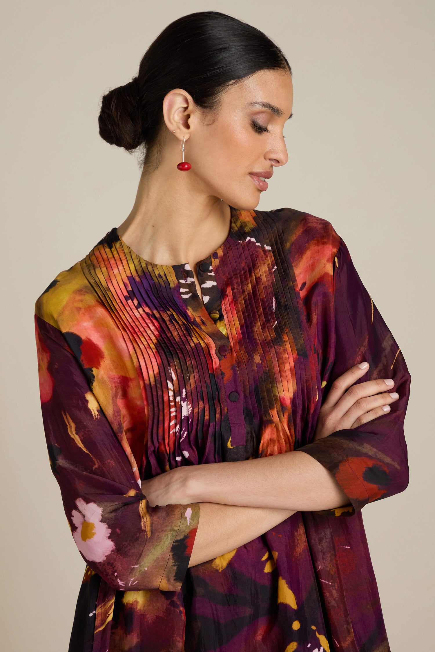 A woman with dark hair in a bun is wearing an Abstract Flower Pleated Dress. She is looking down with her arms crossed, displaying a red earring in one ear.