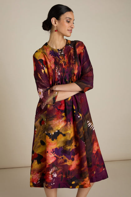 A woman is standing and looking to the side, wearing an Abstract Flower Pleated Dress with an abstract design reminiscent of impressionist art and 3/4 length sleeves. Her dark hair is tied back, and she has red earrings.