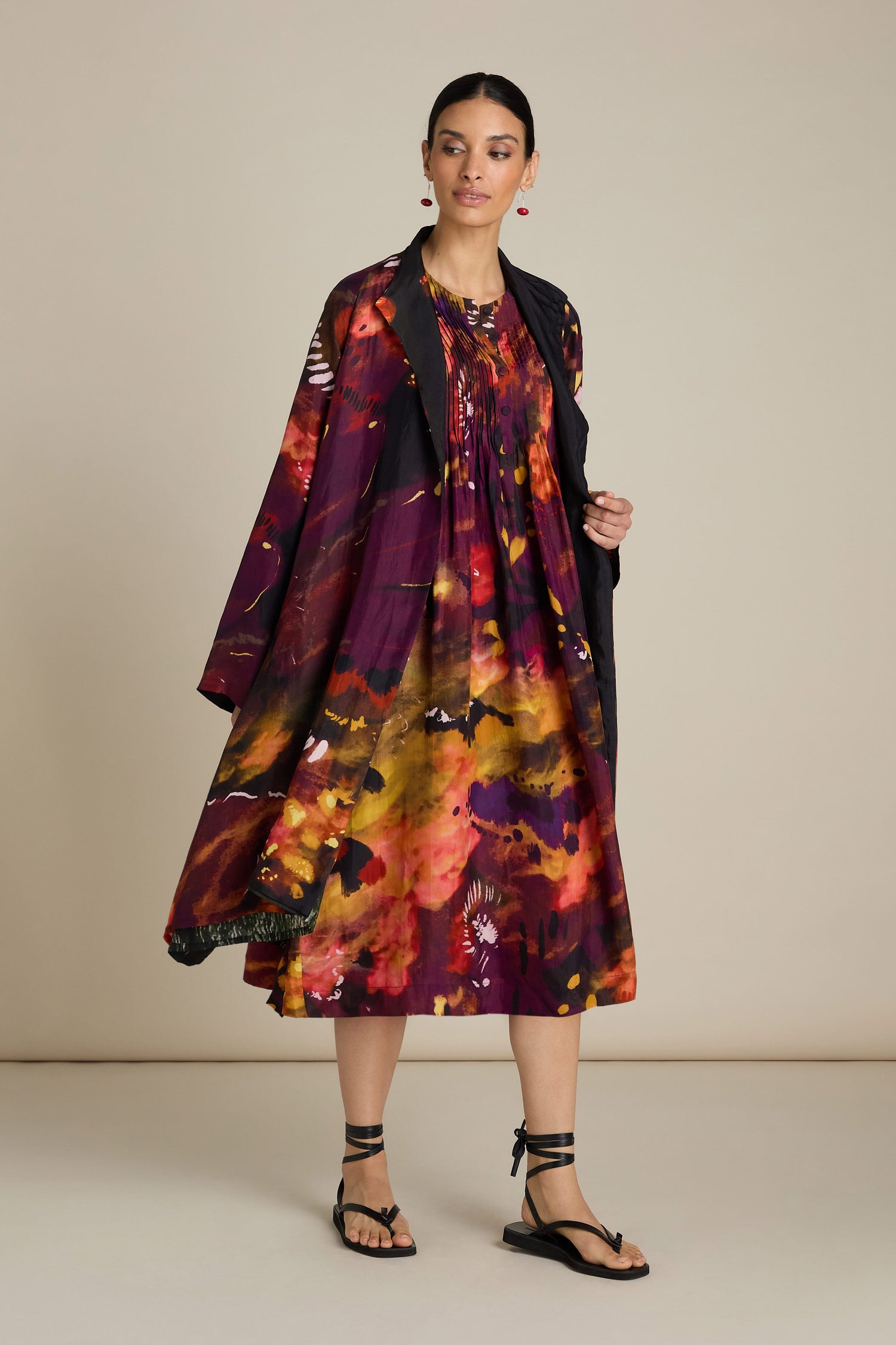 A woman stands against a plain background wearing an Abstract Flower Pleated Dress paired with a matching jacket and black sandals. Her hair is tied back, and she has earrings on, exuding an air of impressionist art in her ensemble.