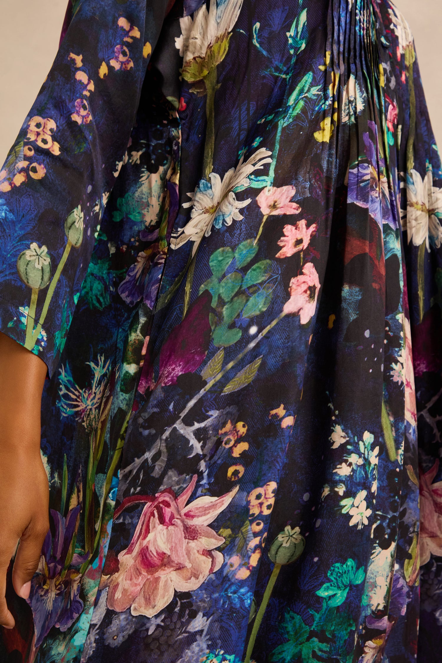 Close-up of the Floral Bouquet Pleated Dress, a dark-colored pleated dress adorned with a vibrant, multicolored floral print and artisan-crafted details. A person's arm is visible on the left side, highlighting the exquisite craftsmanship of this piece.