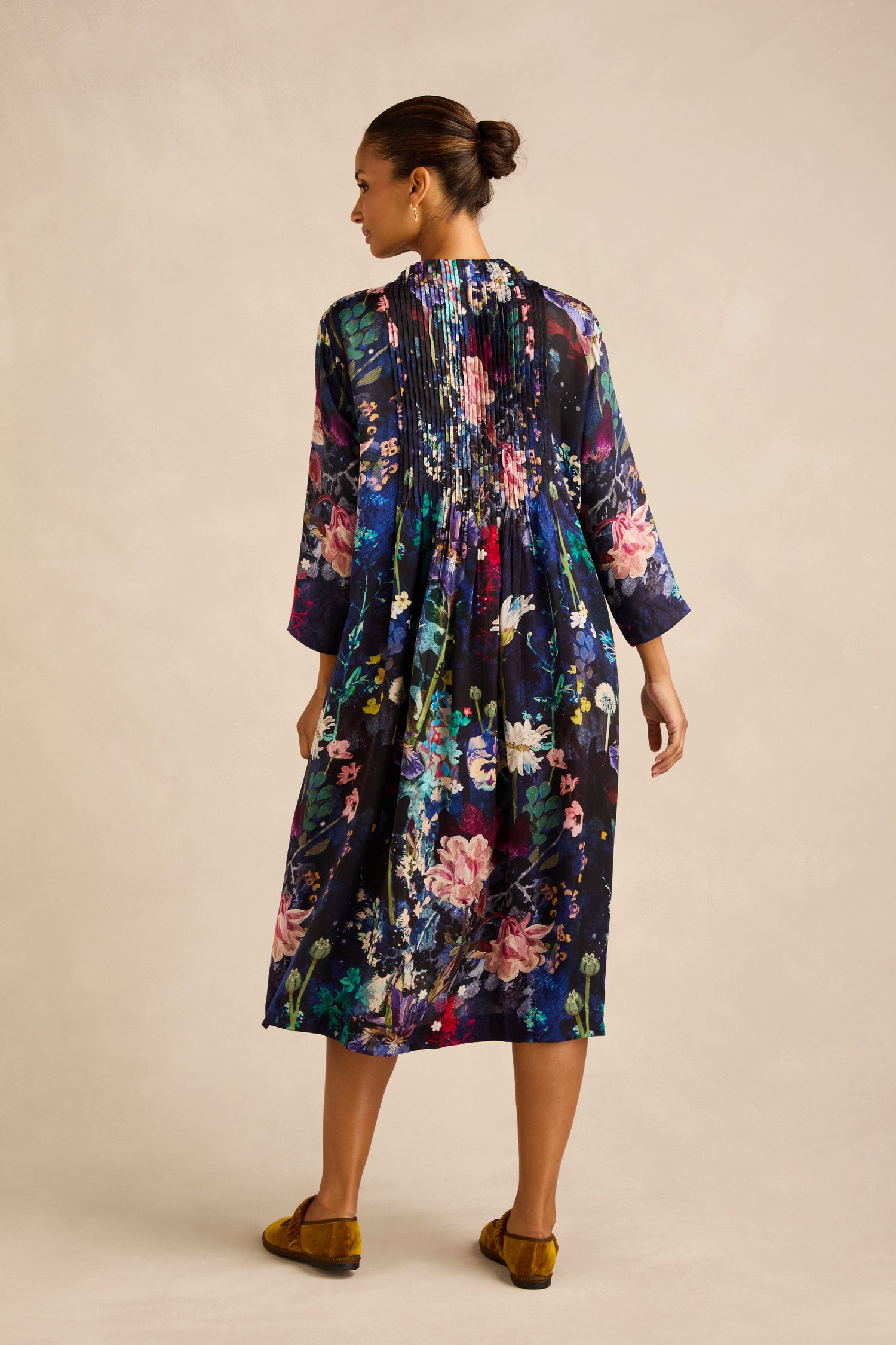 A woman stands facing away, wearing the Floral Bouquet Pleated Dress—a midi-length design crafted from cupro fabric with pleats and three-quarter sleeves—paired with mustard yellow shoes, showcasing beautiful artisan production.
