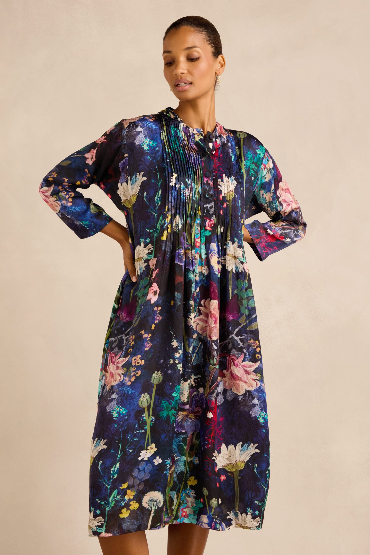 A woman stands with her hands behind her back, wearing the Floral Bouquet Pleated Dress—a loose-fitting, knee-length cupro dress featuring long sleeves and a colorful floral print on a dark background.