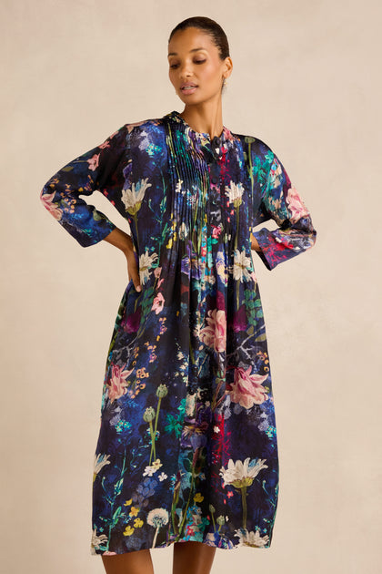 A woman stands with her hands behind her back, wearing the Floral Bouquet Pleated Dress—a loose-fitting, knee-length cupro dress featuring long sleeves and a colorful floral print on a dark background.
