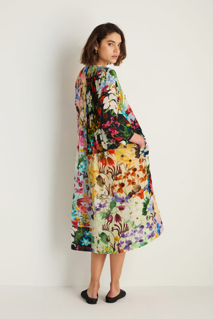 A woman stands against a white wall in the Impressionist Garden Pleated Dress, its colorful floral print and pleated skirt catching the light as she looks over her shoulder in black flat shoes.