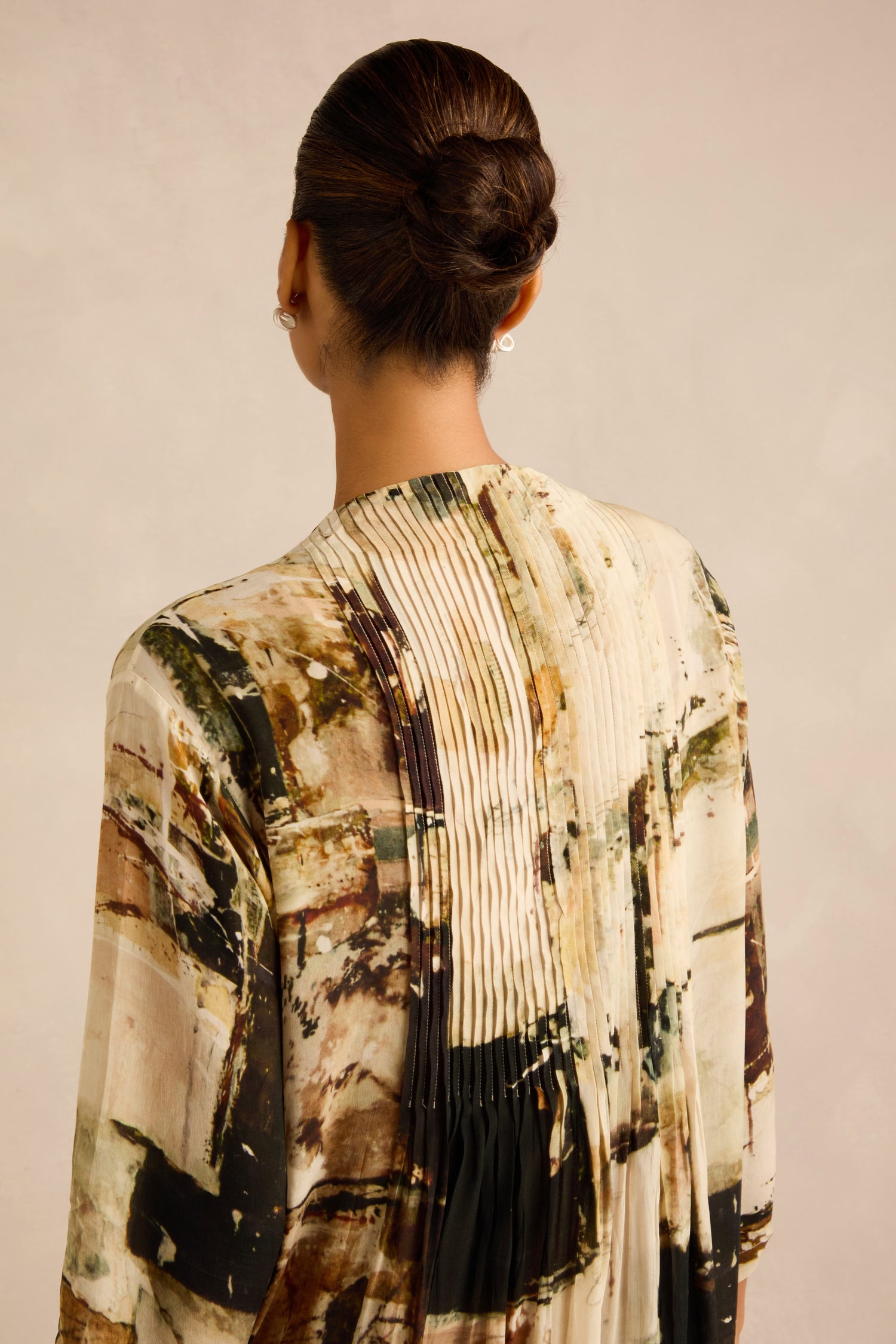 A woman with dark hair in a low bun is shown from behind, wearing a pleated, abstract-patterned dress with earthy tones, similar to the Block Print Pleated Dress.