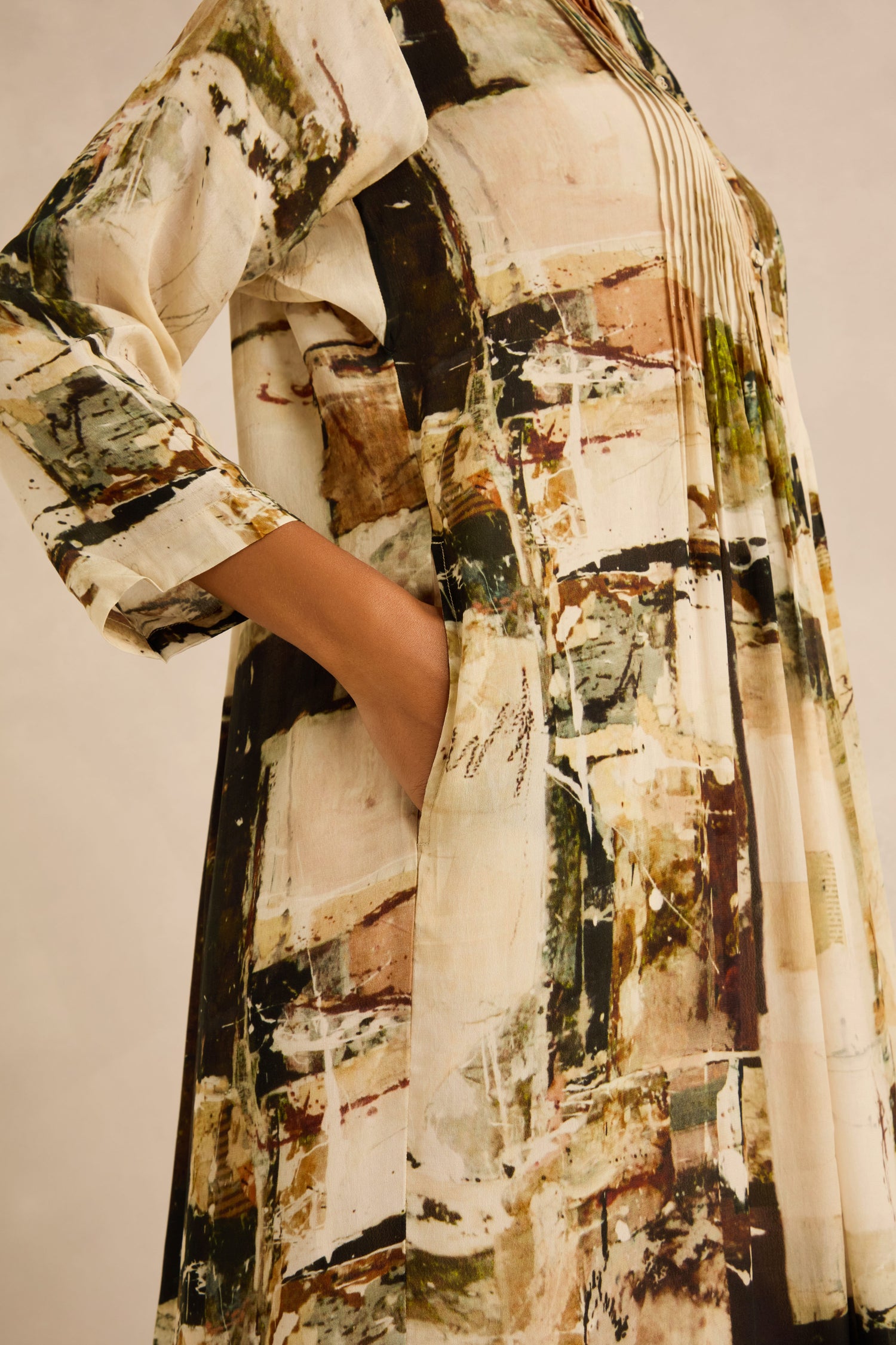 A person wearing the Block Print Pleated Dress, featuring long sleeves and an abstract pattern, with one hand in the side pocket; the background is plain and neutral.