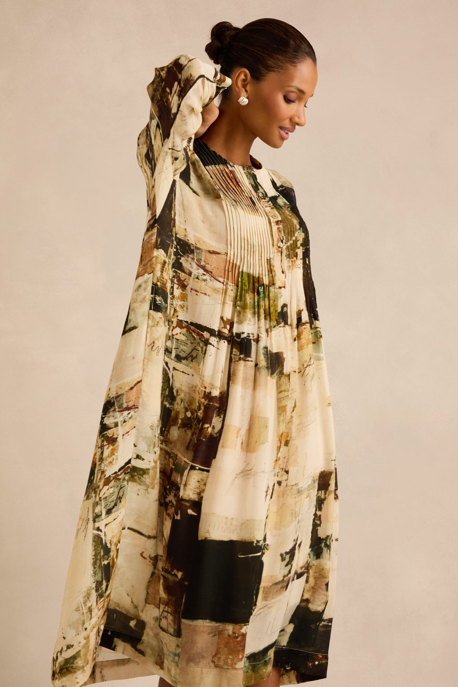 A woman stands sideways against a plain background, one arm raised and smiling slightly, wearing the Block Print Pleated Dress—featuring loose long sleeves and abstract earth-tone patterns.