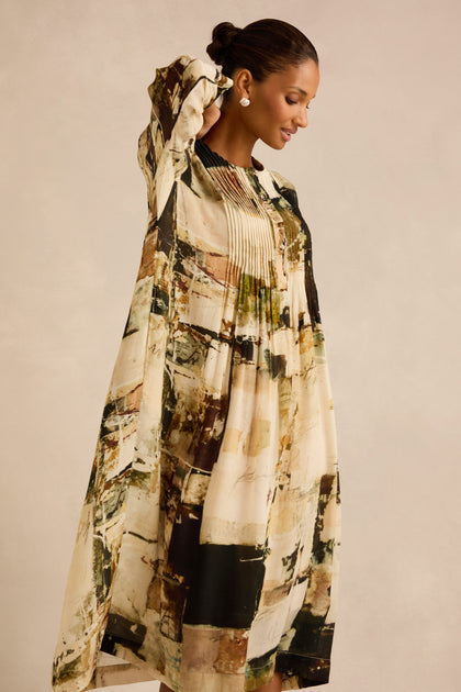A woman stands sideways against a plain background, one arm raised and smiling slightly, wearing the Block Print Pleated Dress—featuring loose long sleeves and abstract earth-tone patterns.