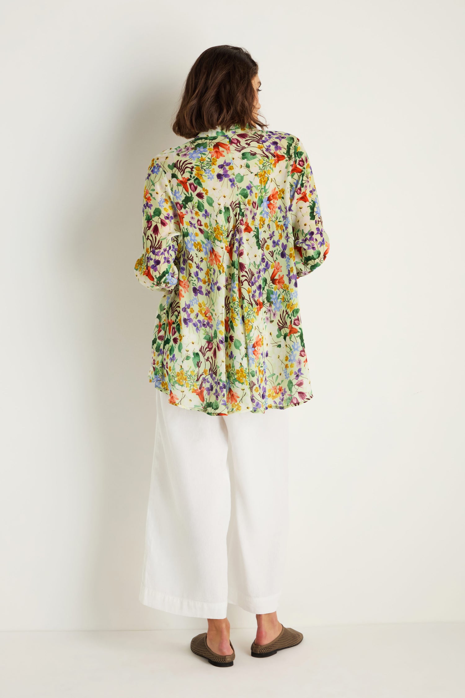 A person faces a white wall, wearing the Country Garden Cupro Shirt featuring an art-inspired floral pattern, paired with white wide-leg pants and brown slip-on shoes.