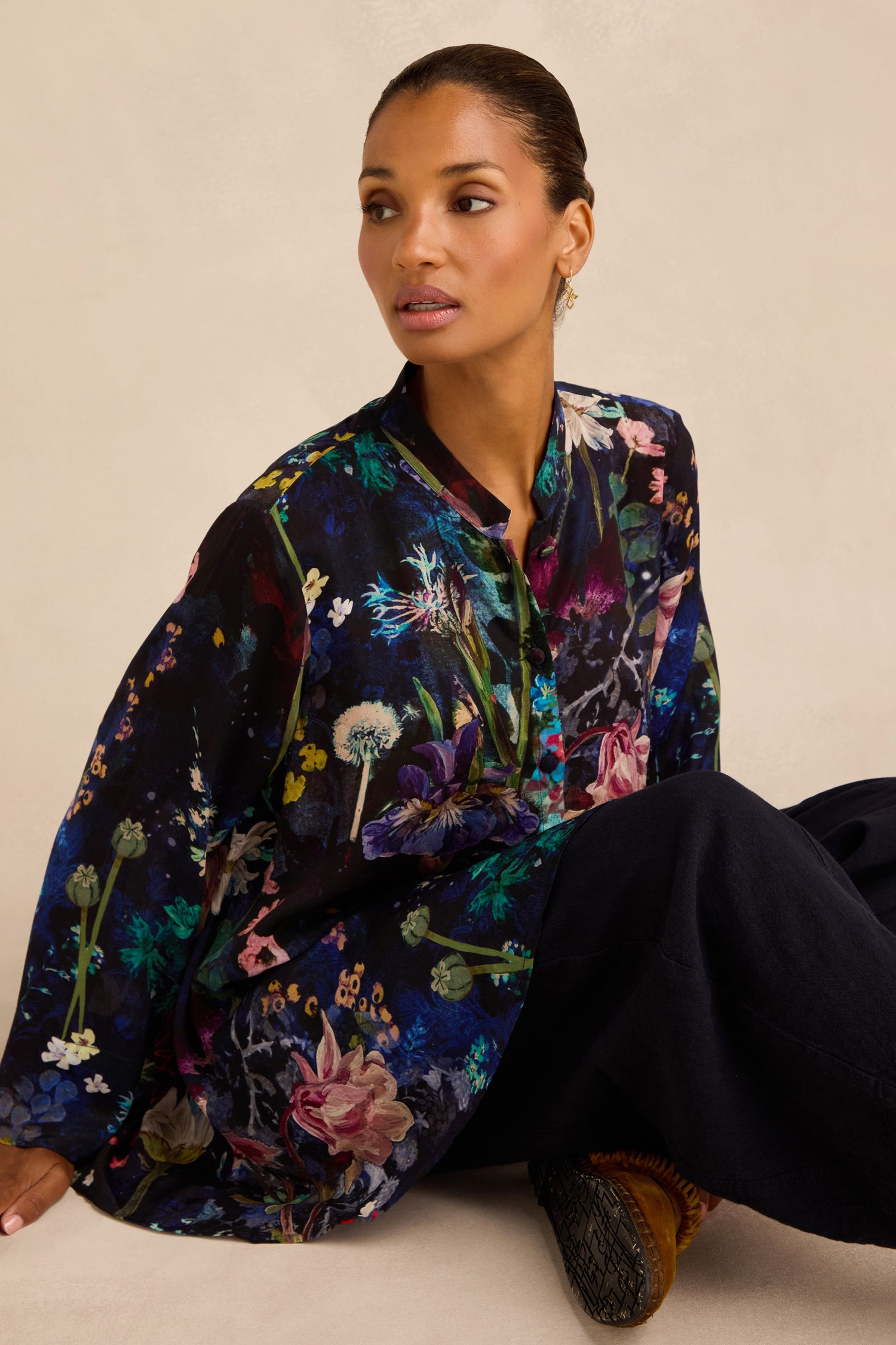 A woman sits on the floor, wearing the Floral Bouquet Flared Shirt— a long-sleeve, button-up top with a dark floral pattern—paired with loose dark pants, looking slightly to the side against a neutral background.