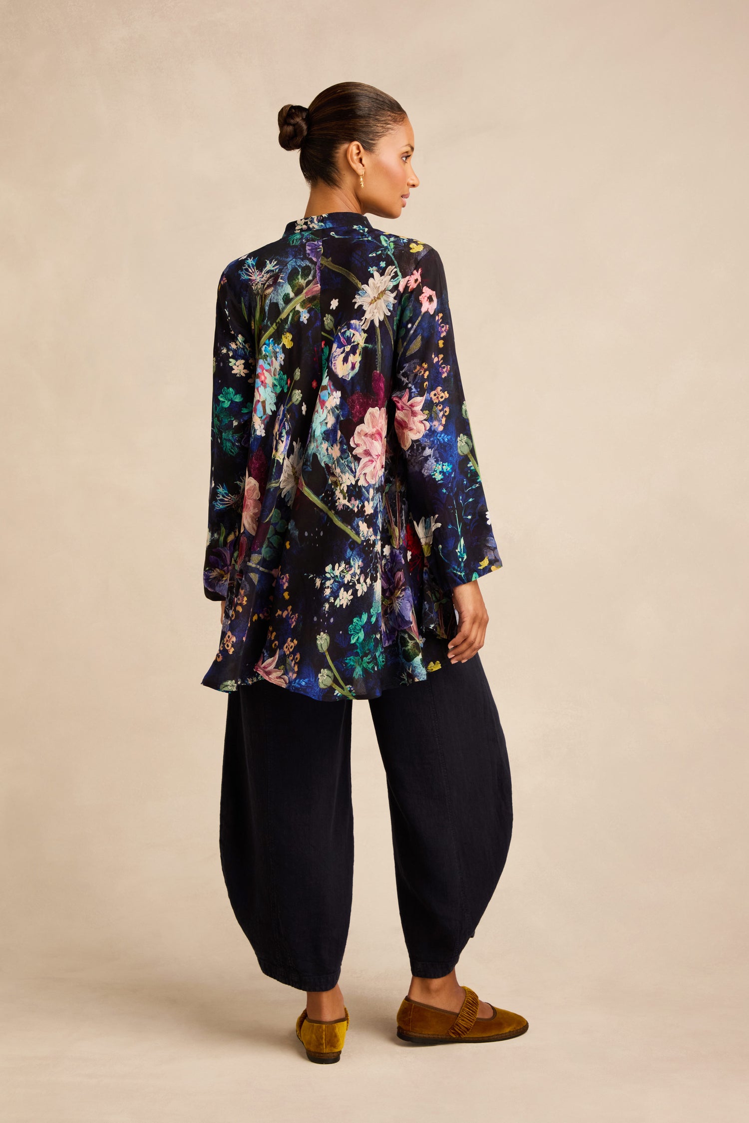 A woman stands facing away, wearing the Floral Bouquet Flared Shirt in a swing silhouette, paired with loose black pants and brown shoes against a neutral background.