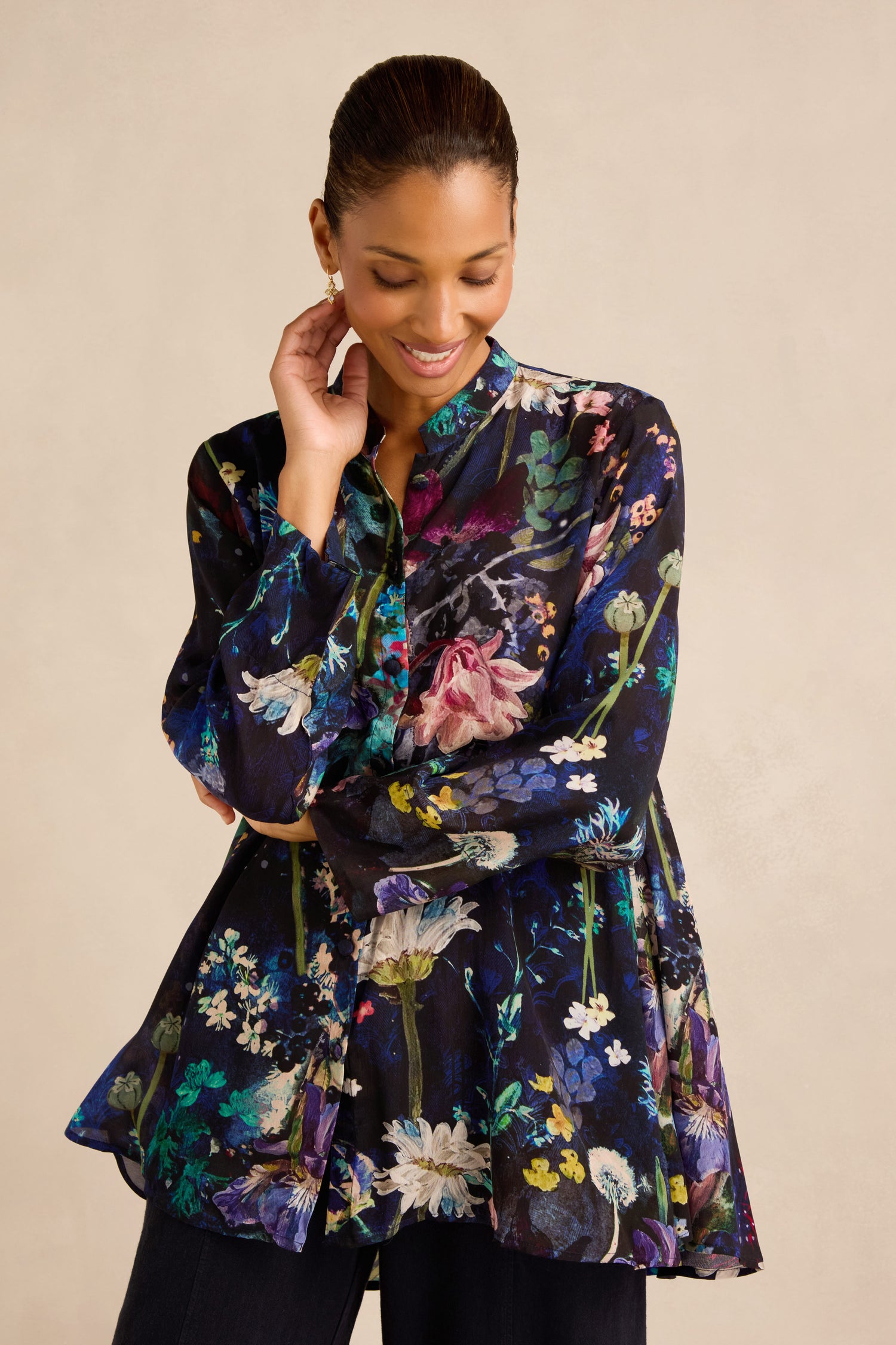 Wearing the Floral Bouquet Flared Shirt, crafted from a soft cupro viscose blend with long sleeves and a charming flared swing silhouette, a woman stands against a plain light background and smiles with her hand touching her face.