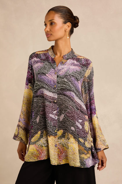 A woman stands against a neutral background wearing the Impressionist Print Flared Shirt—a loose, long-sleeve top crafted from a luxurious cupro and viscose blend with a collar and button front—paired with black pants.