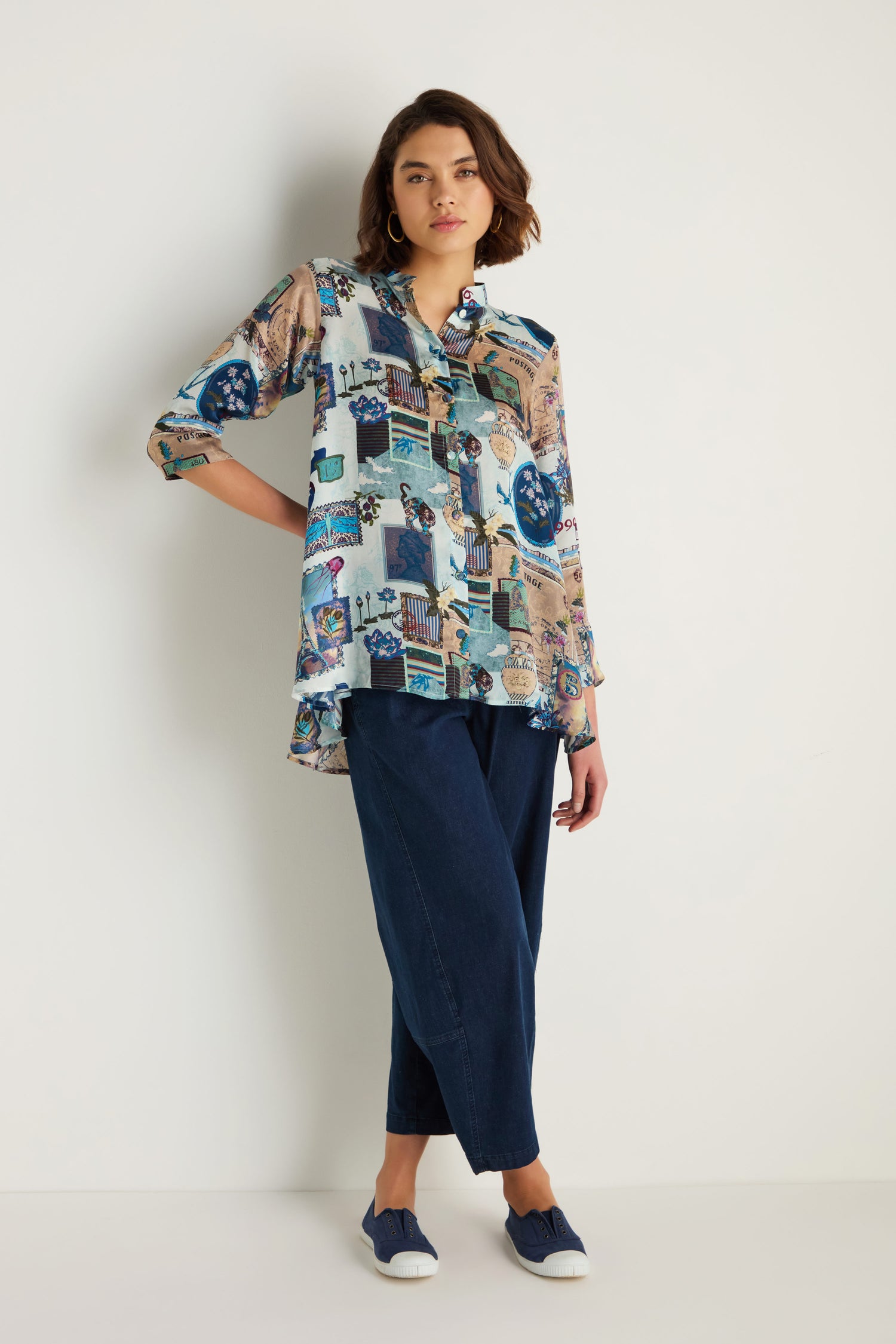 A person stands against a white wall, wearing the Postcard Print Cupro Shirt, loose blue pants that create a relaxed silhouette, and blue slip-on shoes.