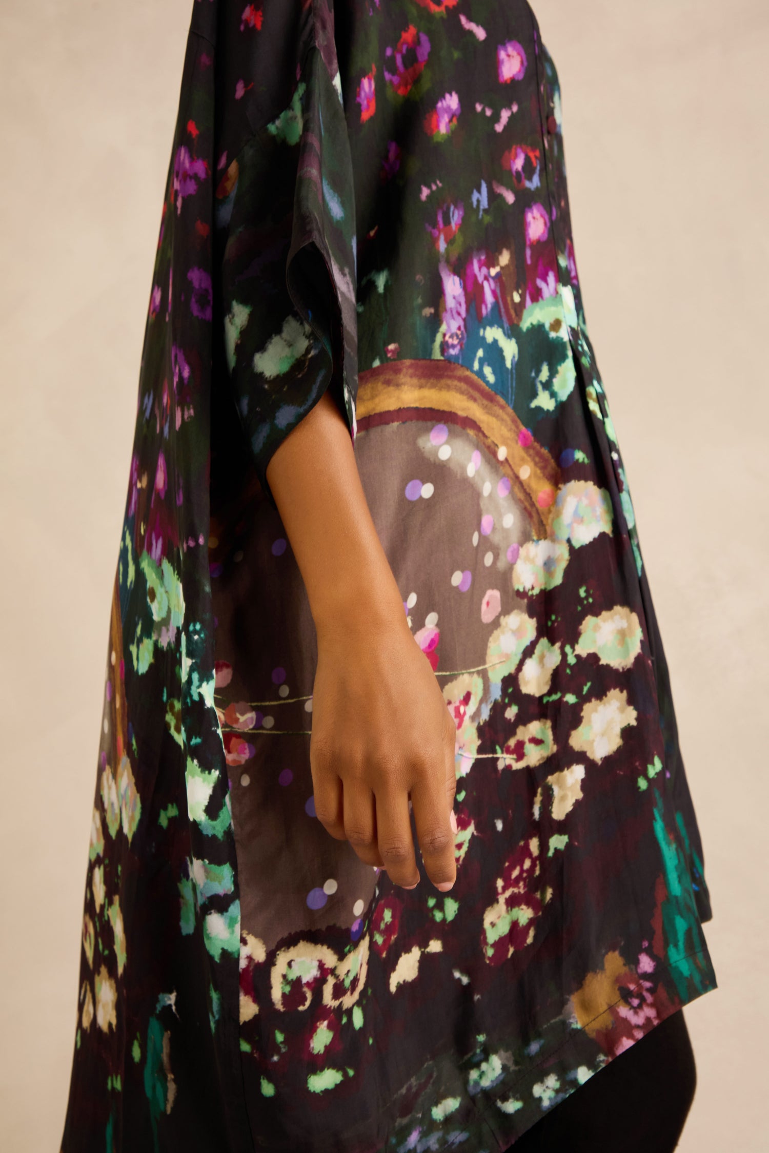 A person wears the Water Lilies Button Through Tunic, a loose-fitting garment adorned with vibrant floral prints in purple, green, and cream accents, standing against a neutral background. This tunic highlights exquisite artisan production in every detail.