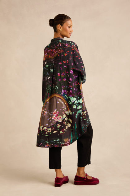 A woman stands sideways against a neutral background, wearing the Water Lilies Button Through Tunic in black with colorful abstract patterns, paired with black pants and red loafers.