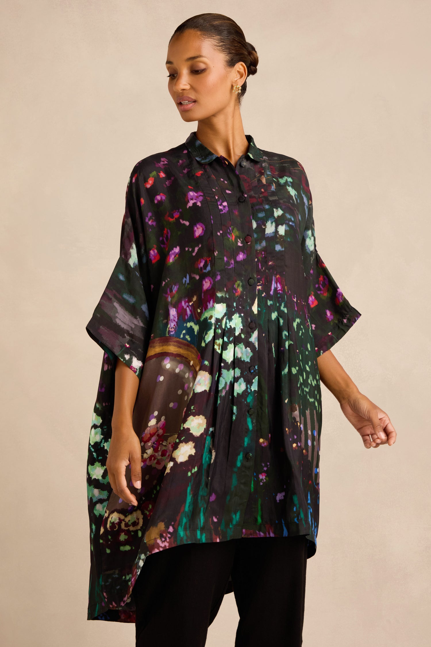 A woman wears the Water Lilies Button Through Tunic, featuring a loose fit and vibrant abstract floral design, paired with black pants and gold hoop earrings, standing against a neutral background—an ideal showcase of artisan production at its finest.