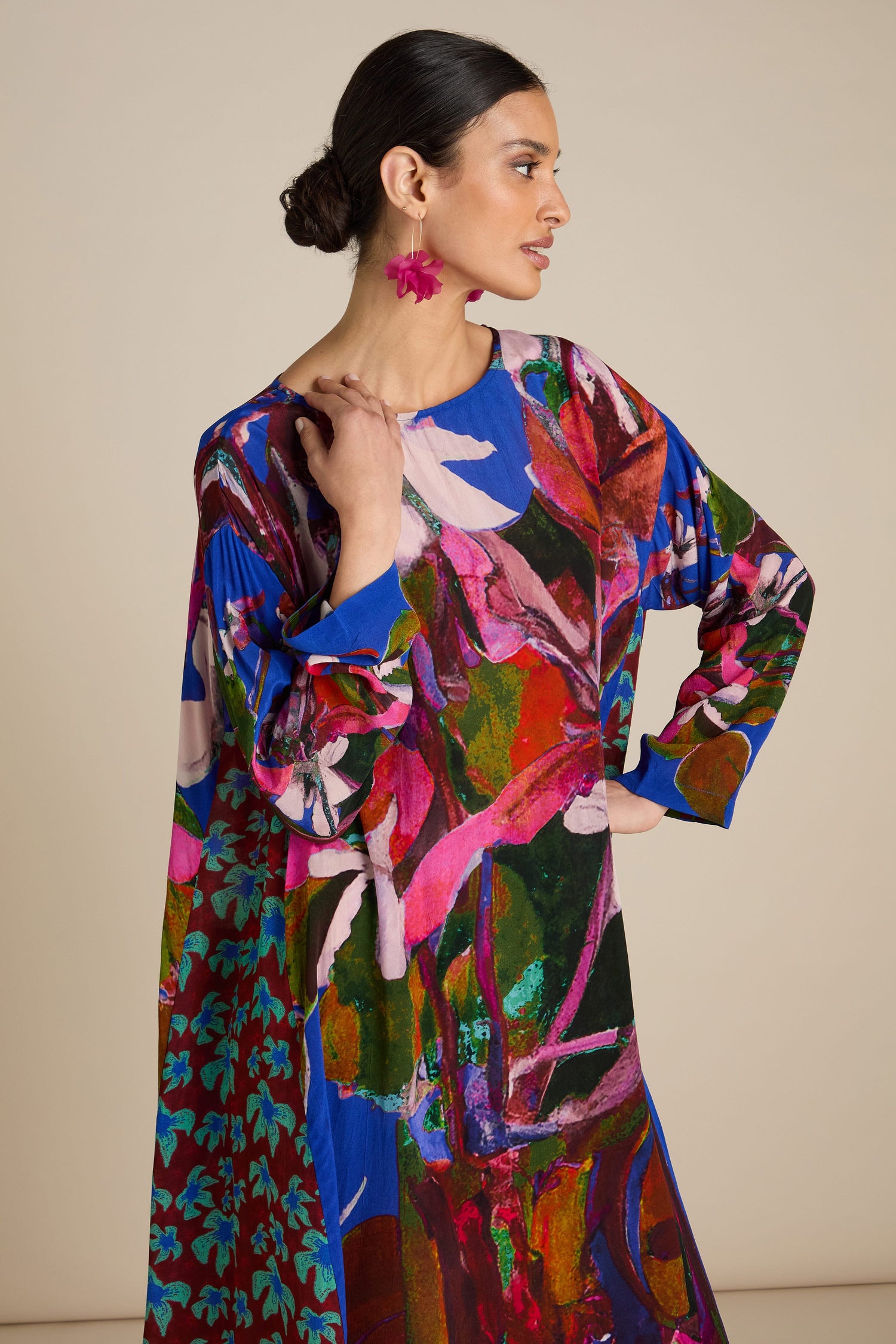 A woman poses wearing a Side Panel Flower Print Dress featuring bold abstract florals. She has her hair in a low bun and is wearing pink flower-shaped earrings, showcasing the relaxed, free-flowing silhouette of her outfit.