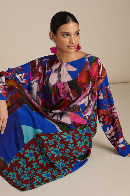 A woman sits in a Side Panel Flower Print Dress, its bold abstract florals and pink floral earrings complementing her relaxed, free-flowing silhouette. She looks to the side with a neutral expression.