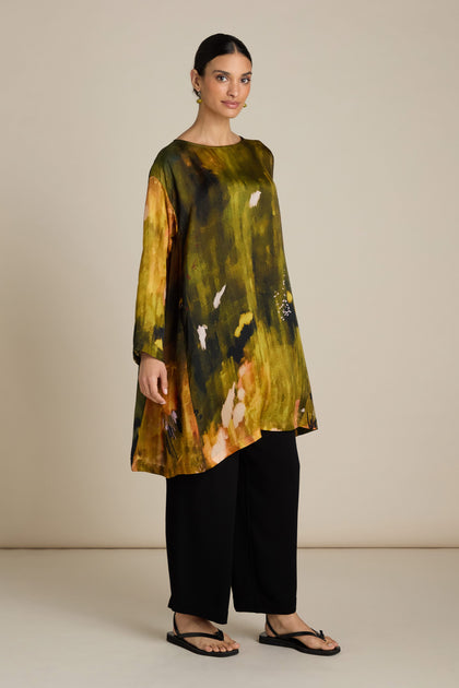 A person stands against a plain background wearing an Autumnal Watercolour Tunic with a relaxed silhouette, paired with black pants and black sandals.