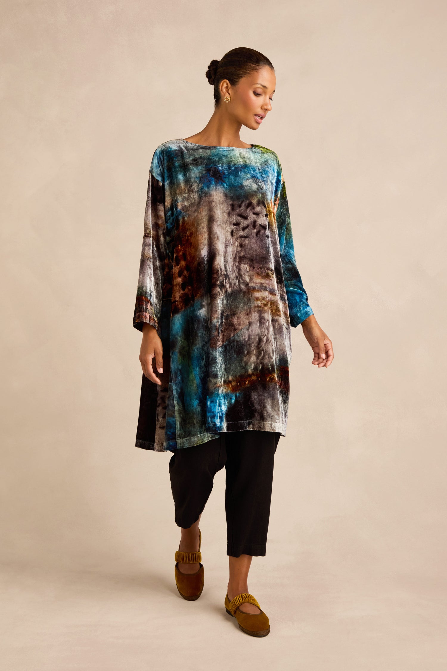 A woman stands against a neutral background, wearing the Sunset Silk Velvet Tunic—a loose, multicolored silk garment with long sleeves—paired with black pants and brown moccasin-style shoes.