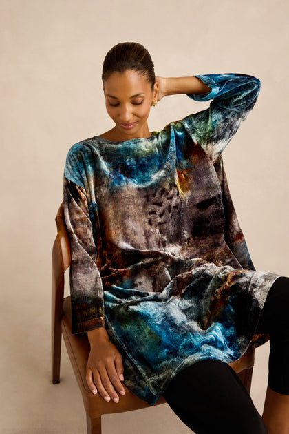 A woman sits on a wooden chair, wearing the Sunset Silk Velvet Tunic with its exclusive multicolored print and long sleeves, paired with black pants, against a neutral background.