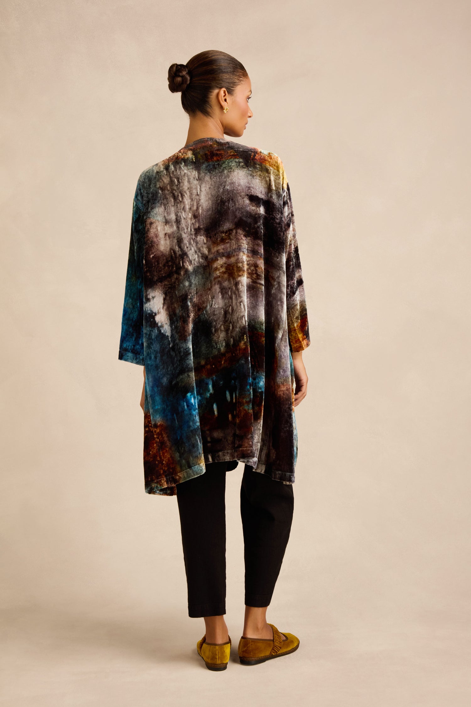 A woman stands facing away, wearing the Sunset Silk Velvet Tunic—a loose, multi-colored jacket featuring an exclusive print—paired with black pants and mustard loafers, her hair styled in a bun against a neutral studio background.