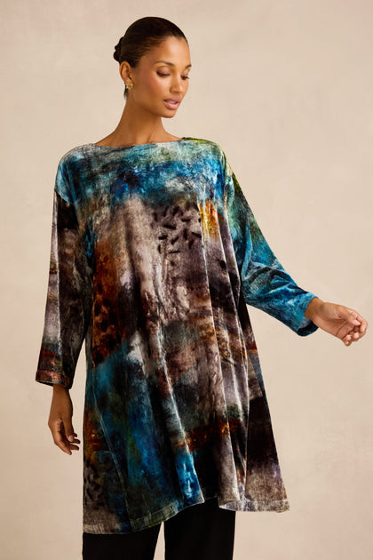 A woman wearing the Sunset Silk Velvet Tunic—a loose, long-sleeved piece featuring an exclusive abstract print in multicolored blue, brown, and black tones—stands against a plain beige background.