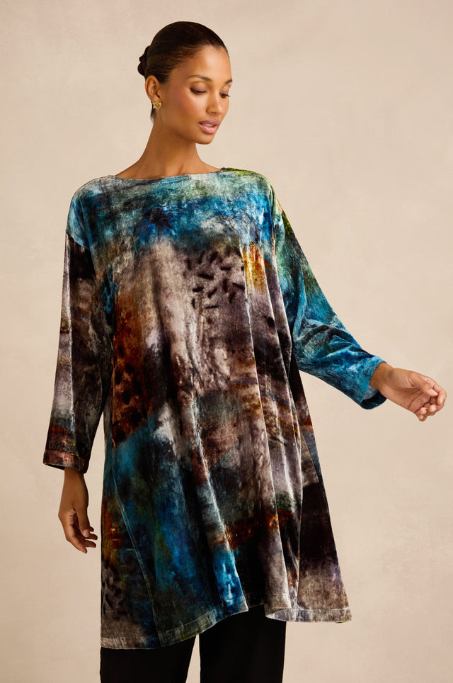 A woman wearing the Sunset Silk Velvet Tunic—a loose, long-sleeved piece featuring an exclusive abstract print in multicolored blue, brown, and black tones—stands against a plain beige background.