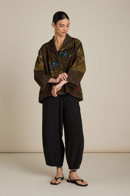 A person standing against a plain background, wearing a Cord Printed Jacket with floral patterns, loose black pants, and black sandals. The 100% cotton brown jacket features a one-button fastening. The person is looking down and has one hand touching their other arm.