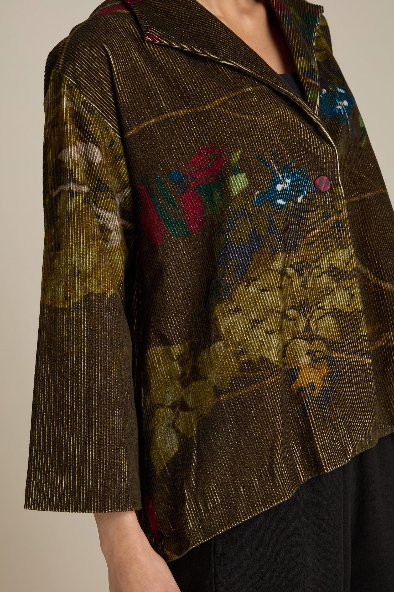 A person wearing a textured, multicolored Cord Printed Jacket with a floral and abstract pattern. The 100% Cotton Cord Printed Jacket features large buttons, including a one-button fastening, a wide collar, and three-quarter length sleeves.