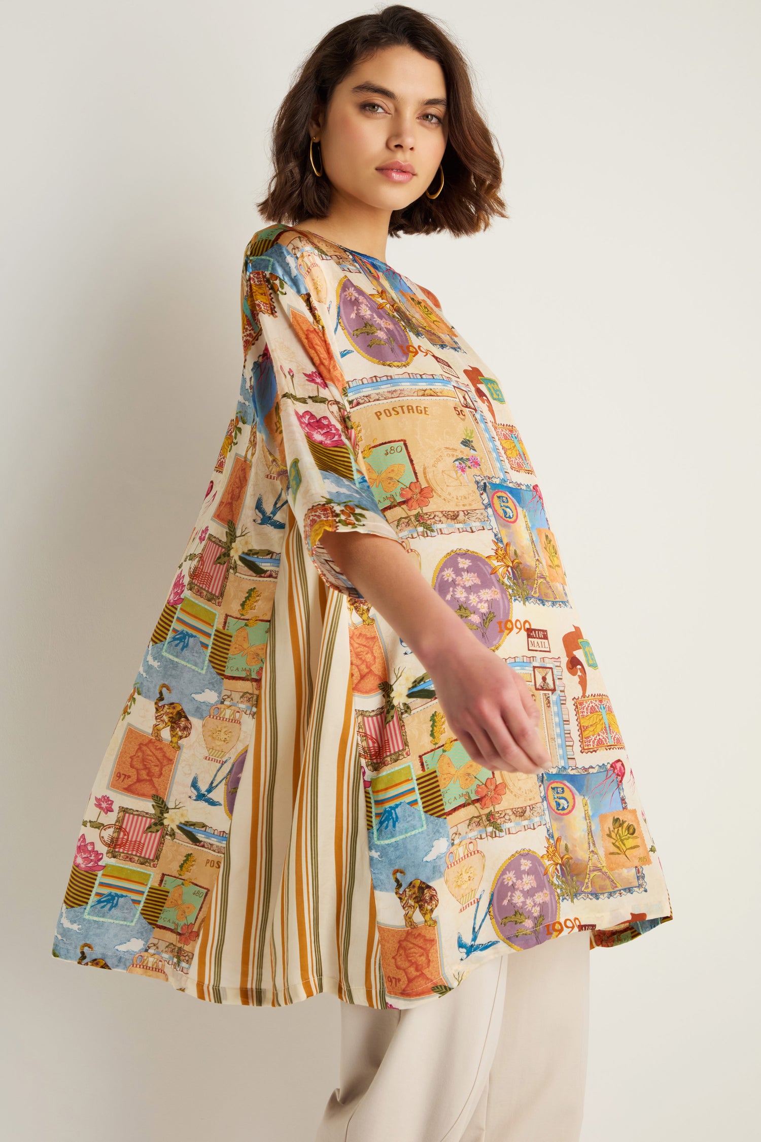 A woman in the Postcard Print Cupro Tunic with hand-drawn postcard and stripe designs, paired with light pants, stands against a plain background.