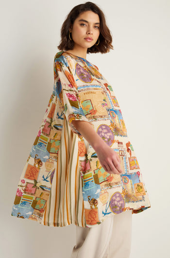 A woman in the Postcard Print Cupro Tunic with hand-drawn postcard and stripe designs, paired with light pants, stands against a plain background.