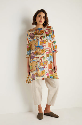 A woman stands against a white wall wearing the Postcard Print Cupro Tunic with vibrant hand-drawn prints, paired with light beige pants and brown slip-on shoes.