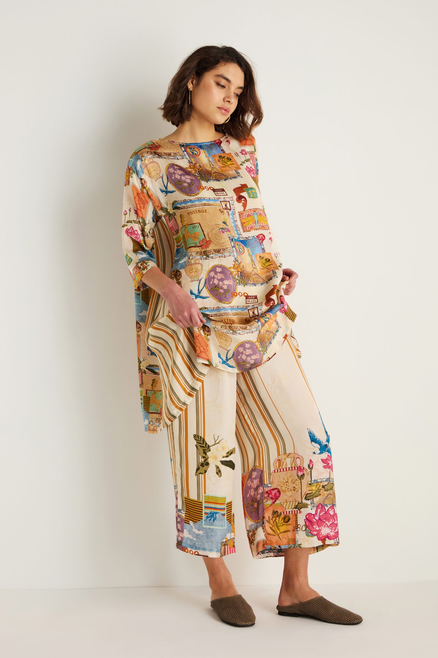 A woman stands against a white wall wearing Postcard Print Stripe Trousers with colorful, eclectic prints inspired by Indian textile artistry, paired with brown slip-on shoes.