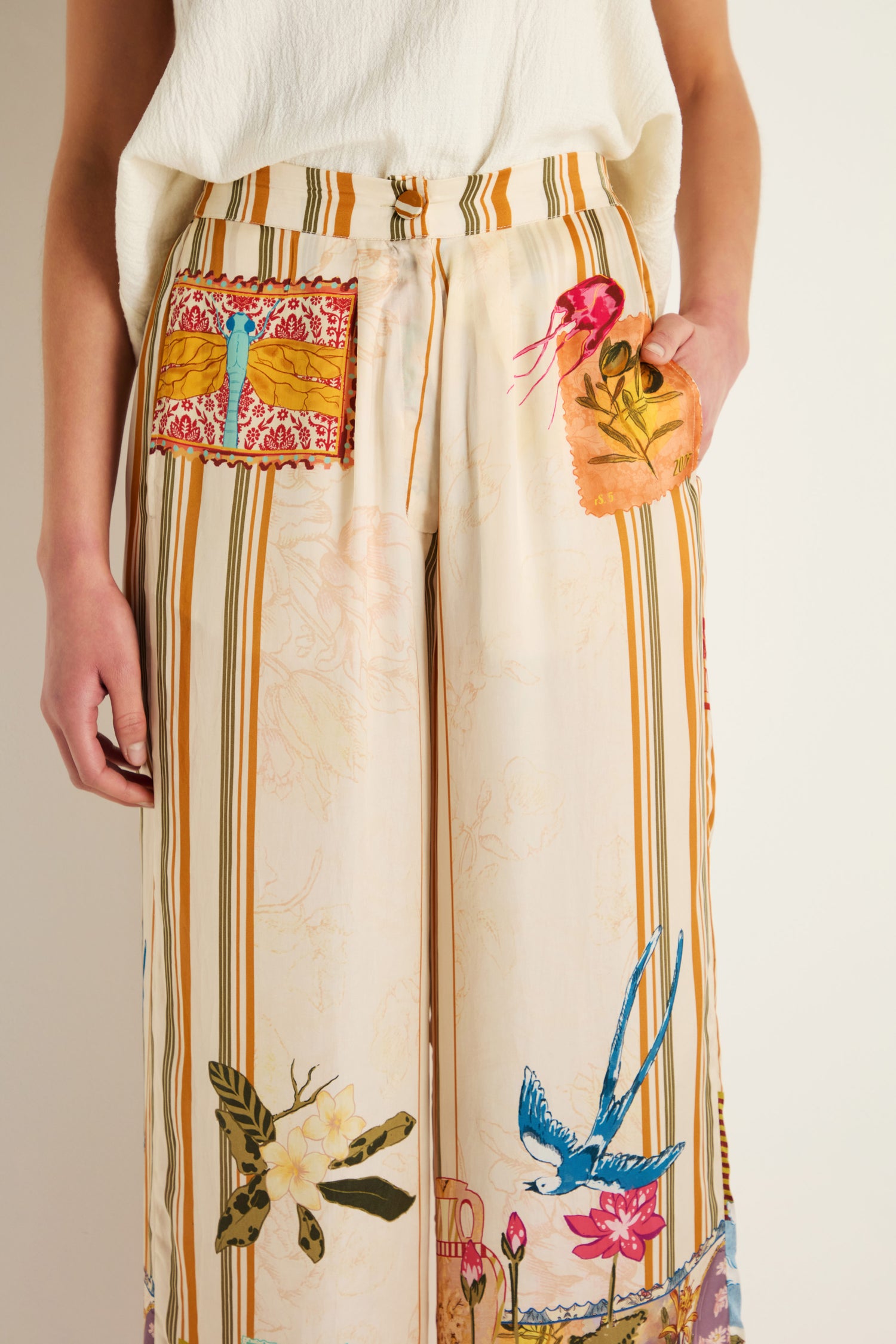 A person wearing Postcard Print Stripe Trousers in cream cupro, featuring vertical stripes and colorful nature-inspired patches like a dragonfly, bird, and floral motifs influenced by Indian textile artistry.