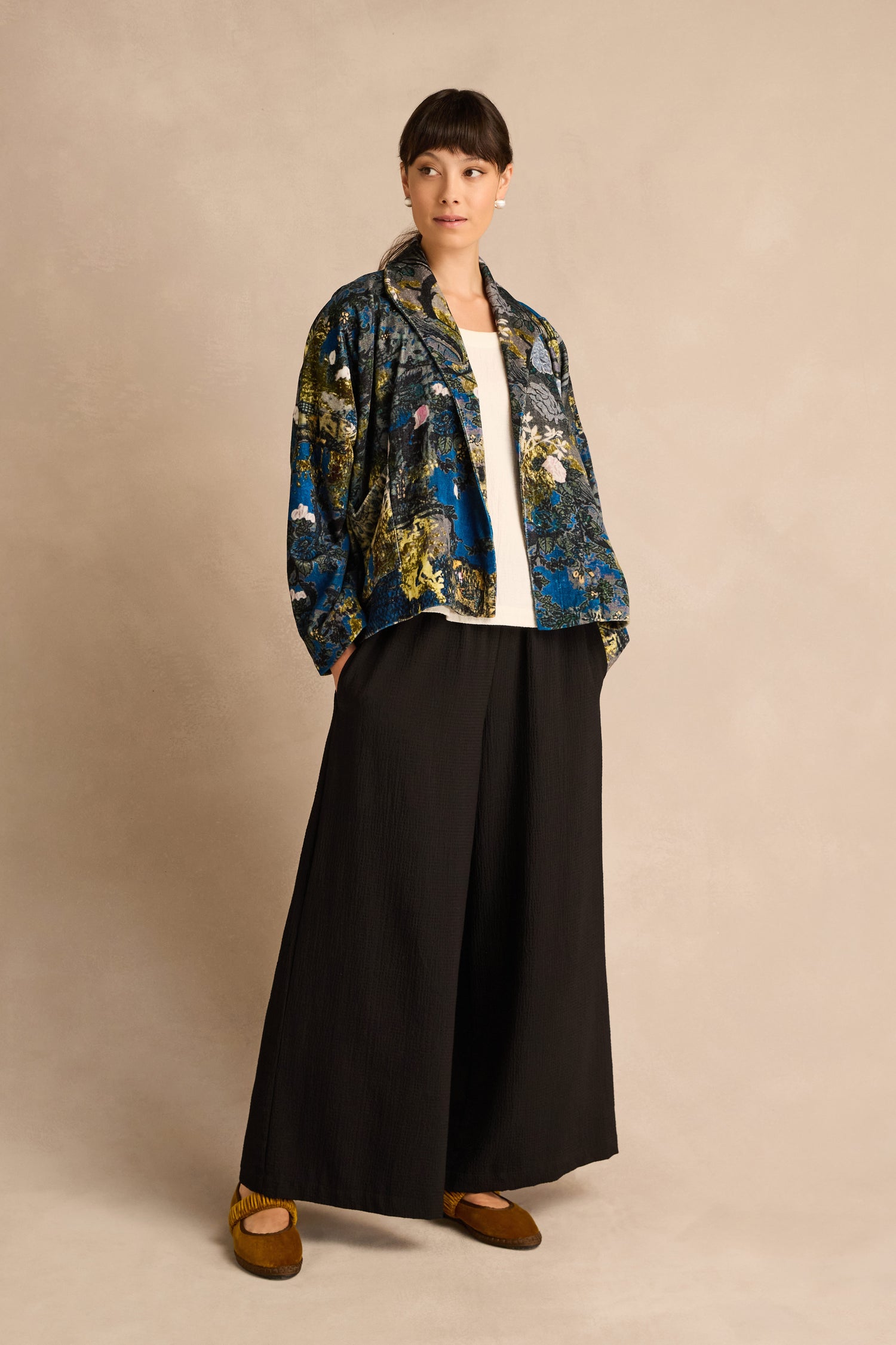 A person stands against a beige background wearing the Japanese Garden Velvet Jacket, paired with a white top, wide black pants, and brown shoes—an ensemble that highlights artisan production and refined elegance.