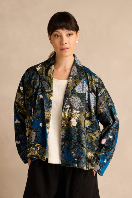 A woman models the Japanese Garden Velvet Jacket, featuring a floral pattern and crafted from artisan-produced cotton velvet, styled over a white top and black pants against a plain beige background.