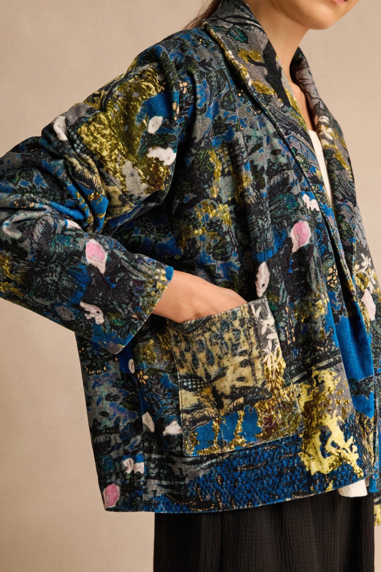 A person, shown from mid-face to hip against a neutral backdrop, wears the Japanese Garden Velvet Jacket—a cotton velvet piece featuring colorful patterns. With a hand tucked into the front pocket, this artisan-made jacket brings a vibrant accent to any outfit.