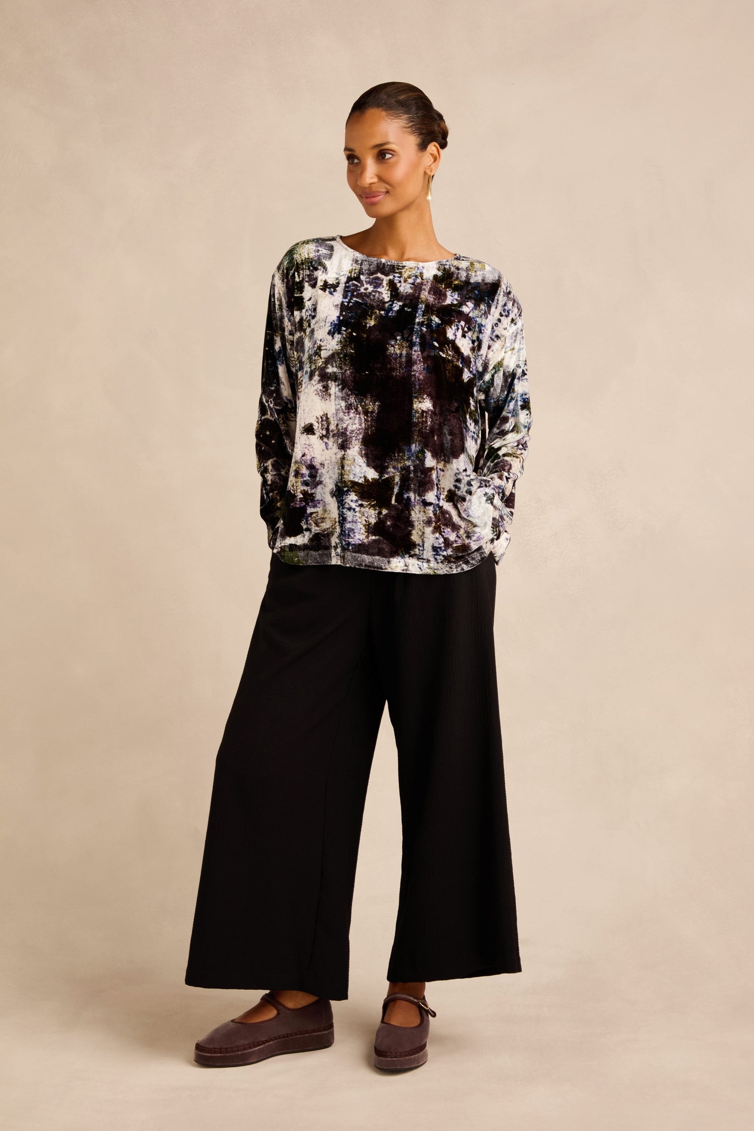 A woman stands against a plain background wearing the Printed Silk Velvet Top, an artisan-made, loose blouse featuring an abstract print and long sleeves. She pairs it with wide black pants and dark flat shoes. With her hands in her pockets, she gazes slightly to the side.