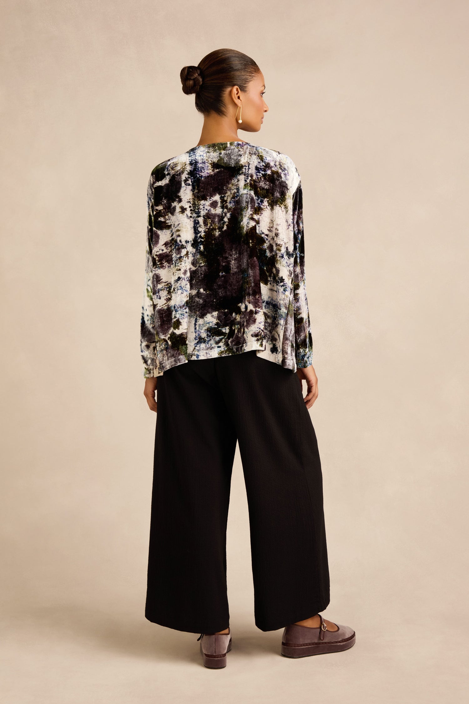 A person stands facing away, wearing the Printed Silk Velvet Top with wide black pants and platform shoes against a plain, neutral background.