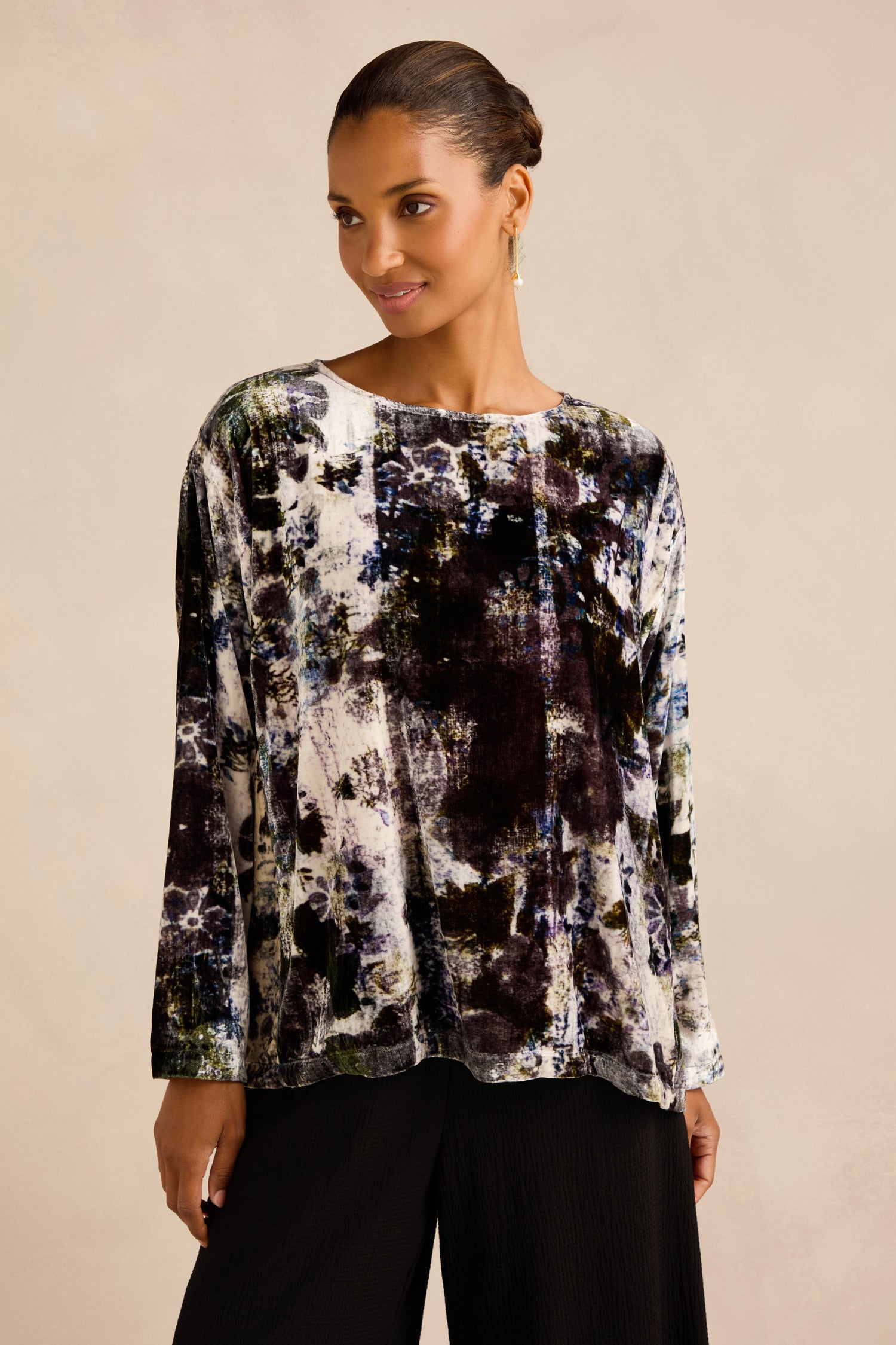 A woman stands against a plain beige background wearing the Printed Silk Velvet Top—an artisan-made, long-sleeve blouse featuring an abstract print in black, white, and blue tones—paired with black pants.