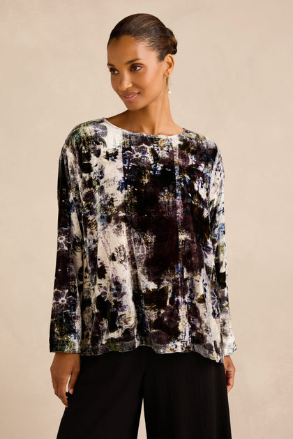 A woman stands against a plain beige background wearing the Printed Silk Velvet Top—an artisan-made, long-sleeve blouse featuring an abstract print in black, white, and blue tones—paired with black pants.
