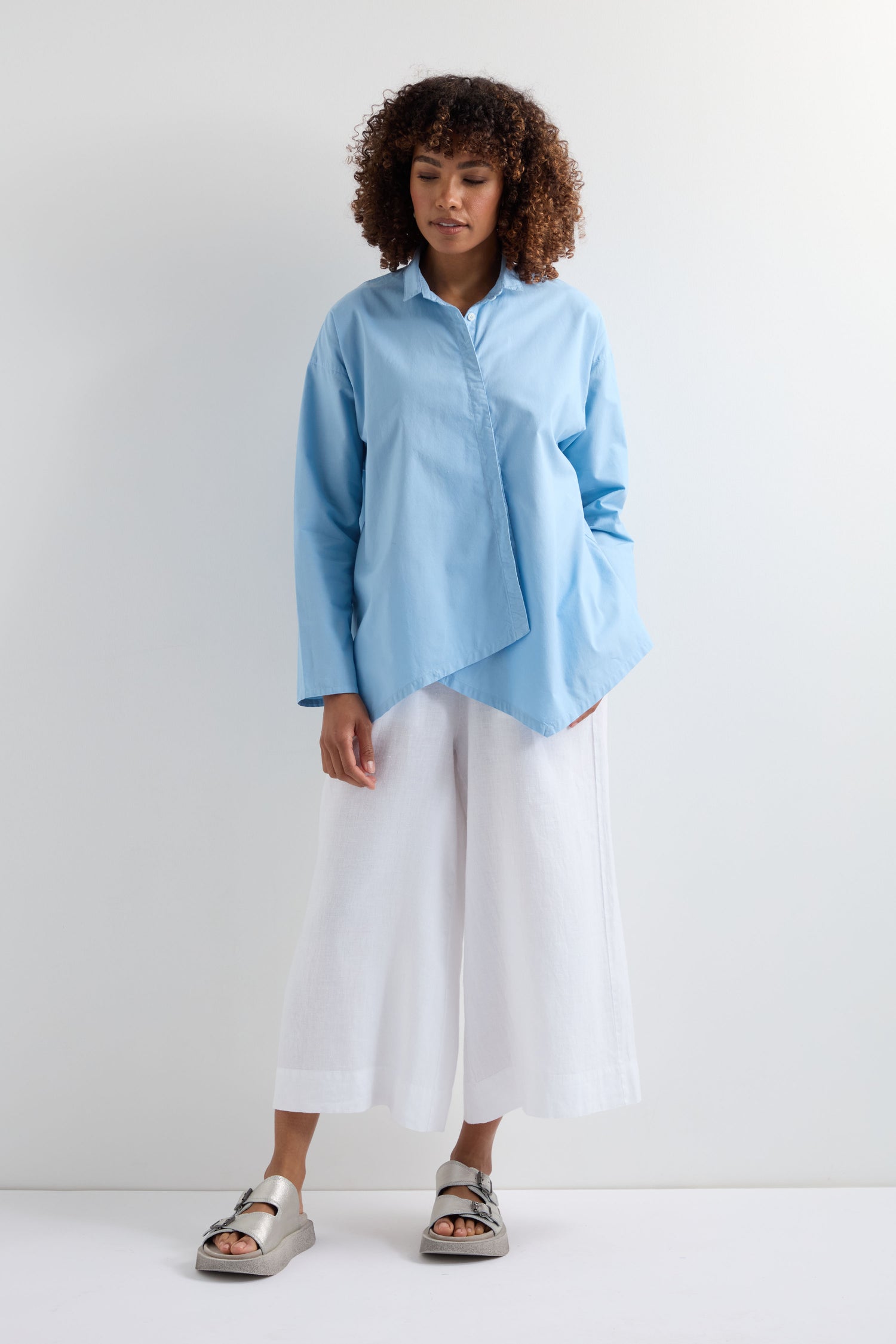 A person stands before a plain white wall, dressed in a light blue Saifu Shirt with relaxed tailoring, wide white pants, and light-colored sandals.