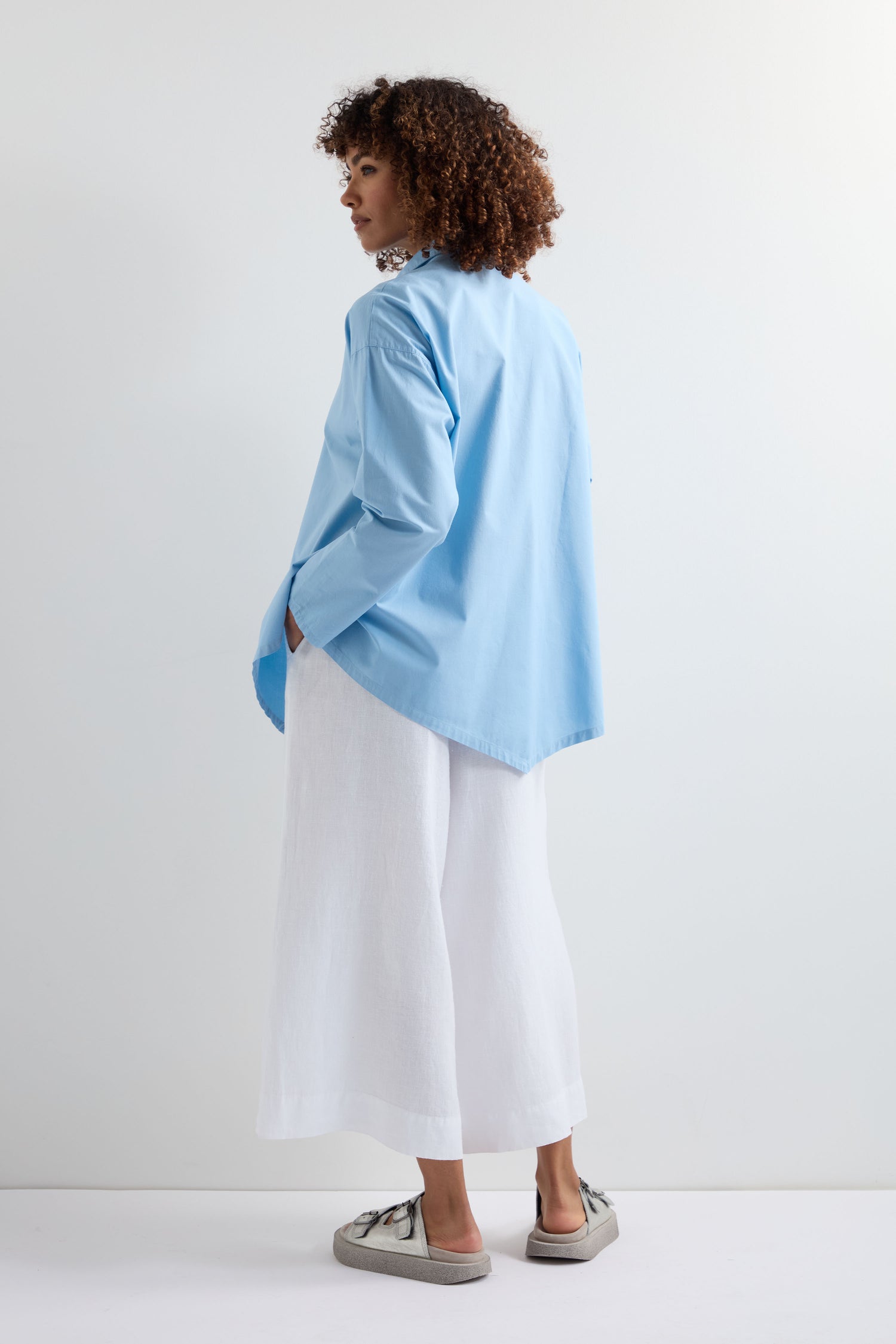 A person stands facing away, wearing a long light blue Saifu Shirt with relaxed tailoring, loose white pants, and gray sneakers against a plain white background.