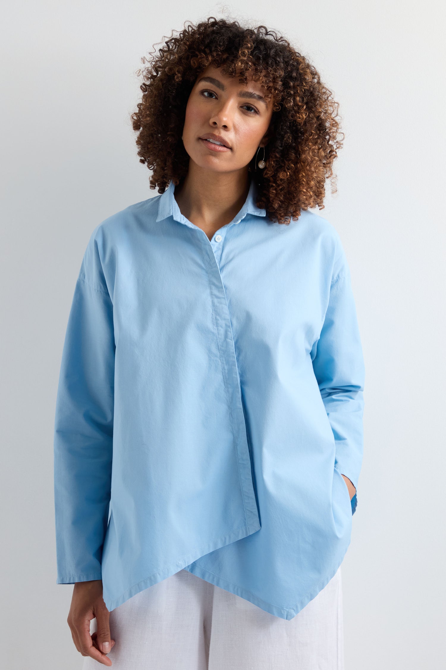 A person with curly hair wears the Saifu Shirt, known for its relaxed tailoring and asymmetrical front, paired with white pants against a plain light background.