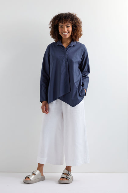 A person stands against a plain background, wearing a navy blue Saifu Shirt with relaxed tailoring, paired with white wide-leg pants and light-colored sandals.