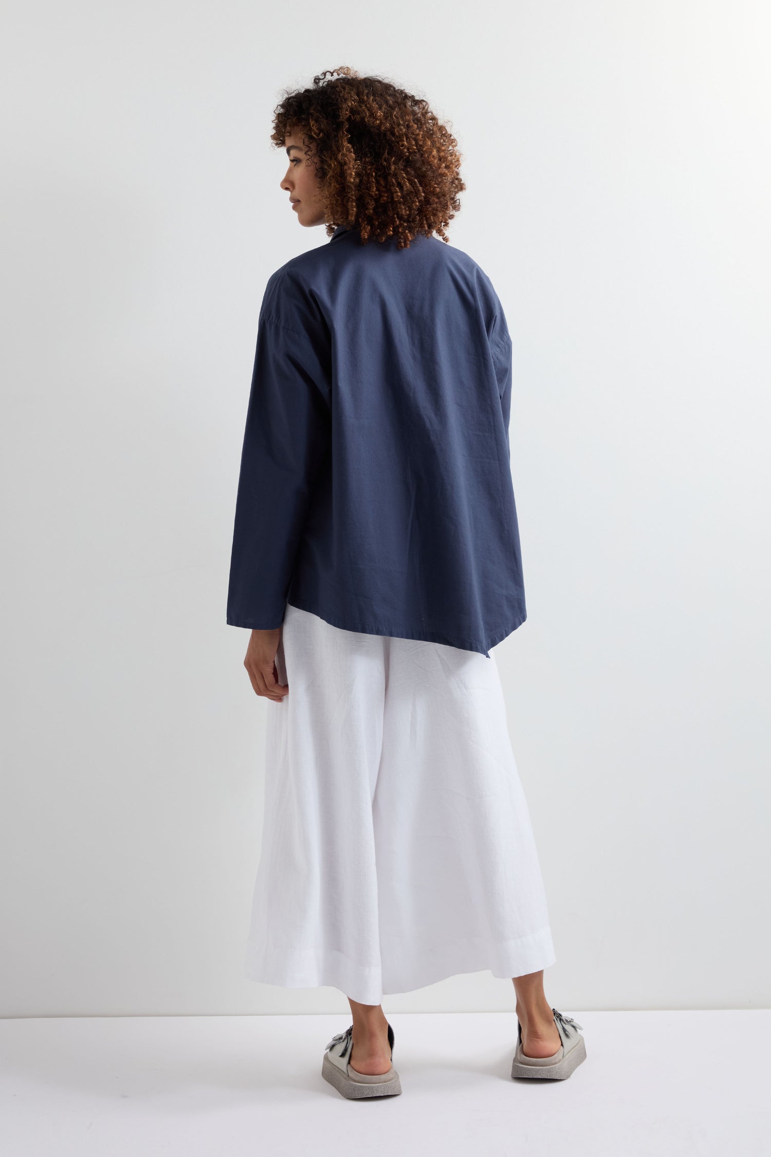 A person with curly hair faces away, wearing a navy Saifu Shirt with relaxed tailoring, white wide-leg pants, and gray shoes against a plain white background.
