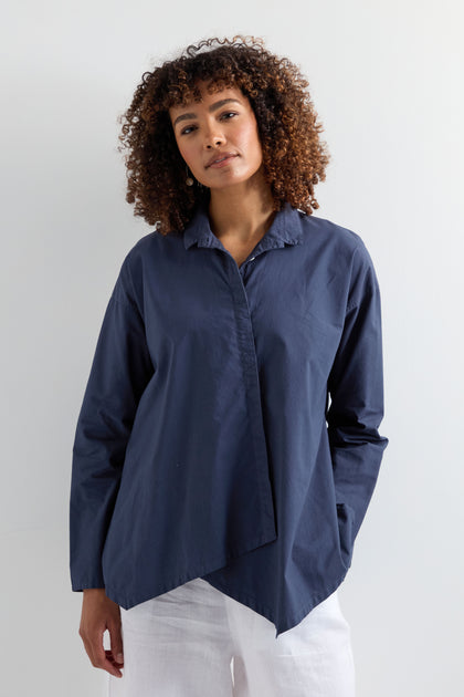 A person with curly hair wears a dark blue Saifu Shirt with relaxed tailoring and white pants, standing against a plain light background.