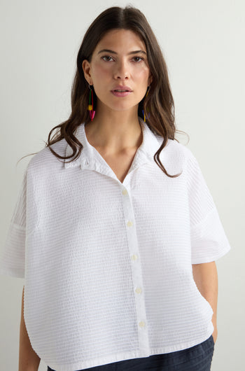 A woman with long brown hair models a Pleated Short Boxy Shirt made of luxurious cotton lawn, paired with colorful dangly earrings, set against a plain light background.